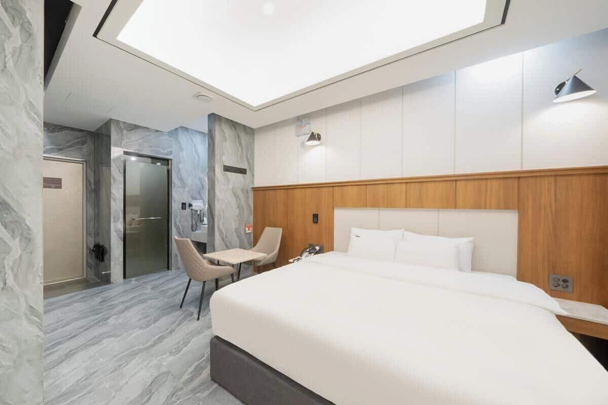 Ein modernes Hotelzimmer mit Marmorboden, einem Holzkopfende, einem Bett, einer Sitzecke und einer Glastür zum Badezimmer.