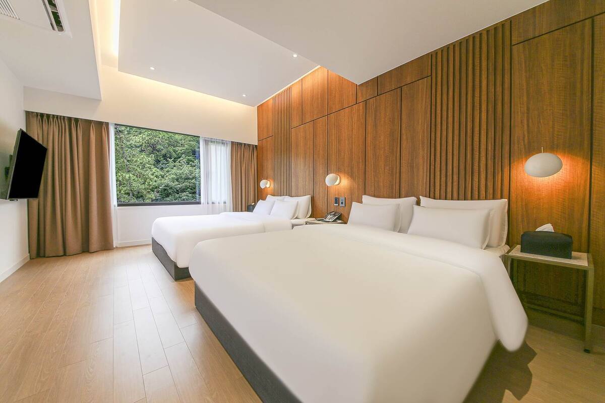 Ein modernes Hotelzimmer mit zwei Betten, einem großen Fenster mit Vorhängen, Holzvertäfelung an den Wänden und einem wandmontierten Flachbildfernseher.