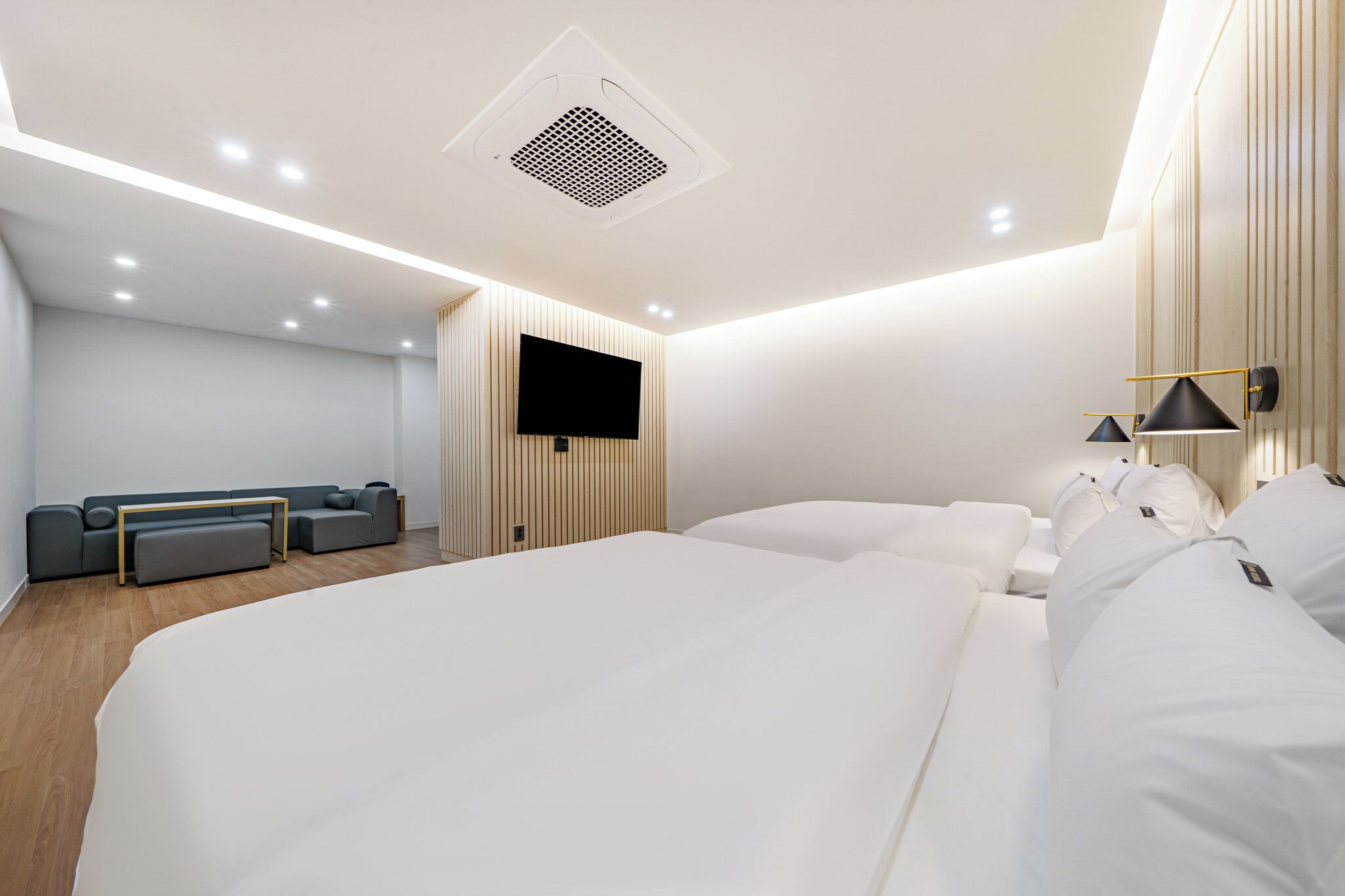 A modern hotel room with a large bed, a seating area with a sofa and ottoman, a flat-screen TV mounted on a wooden panel wall, and a ceiling with recessed lighting.
