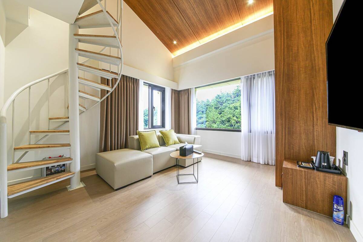 Ein modernes Wohnzimmer mit Wendeltreppe, einer Sofagarnitur, einem Couchtisch und einem großen Fenster mit Blick auf Grünflächen.