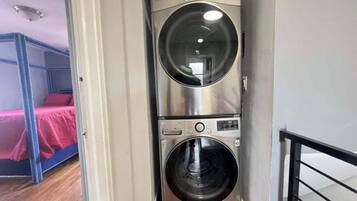 Un espace buanderie compact comprenant deux machines à laver superposées, un lit superposé avec une couverture rouge et une rambarde en métal noir.