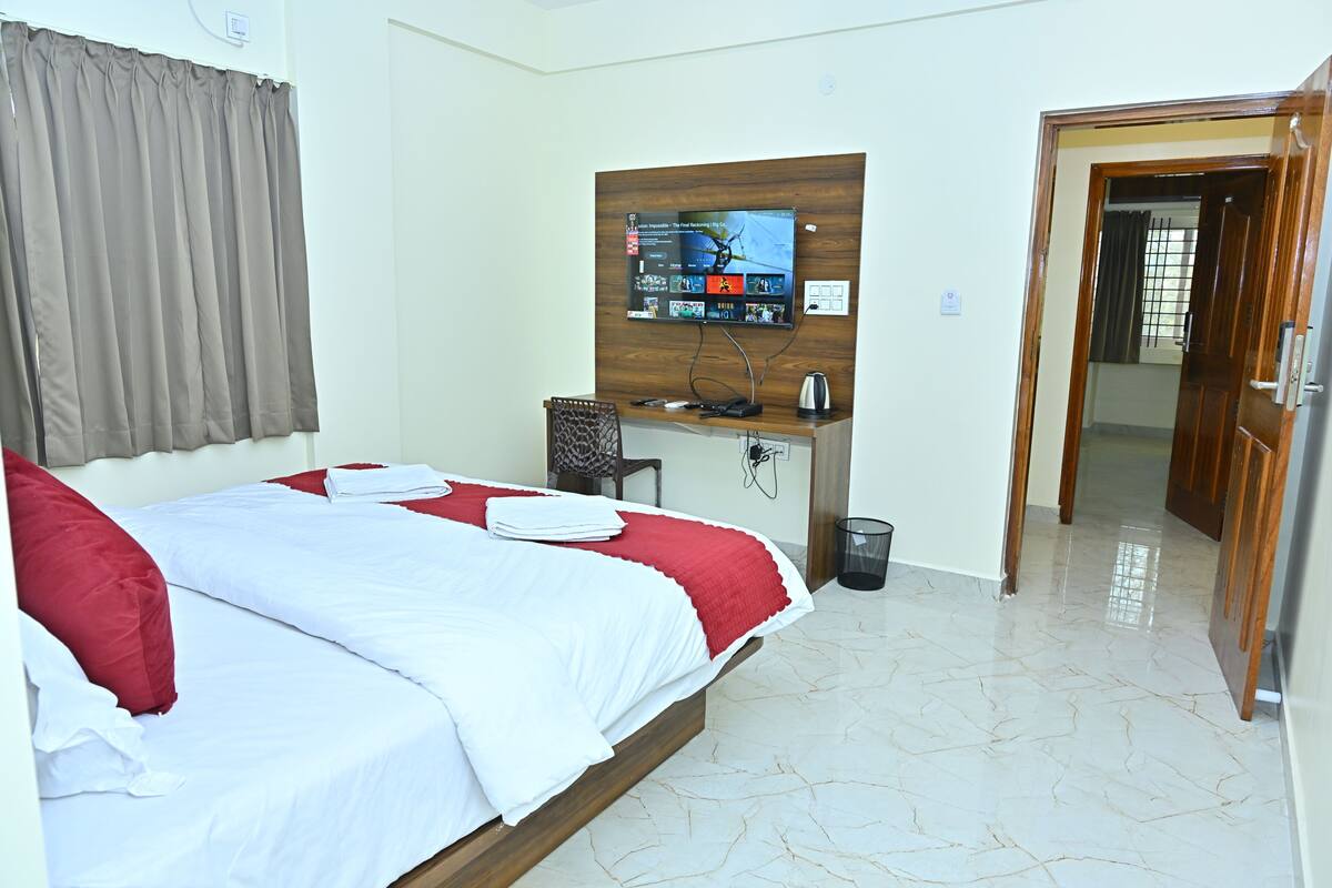 Habitación de hotel con una cama, un escritorio con computadora, un televisor montado en la pared y una puerta que da a otra habitación.