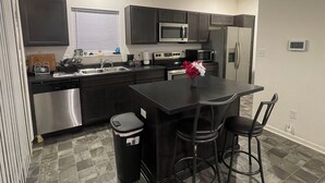 A modern kitchen with dark cabinets, stainless steel appliances, a central island with bar stools, and a trash can. - 3-bedroom townhome with AC (Indianapolis)