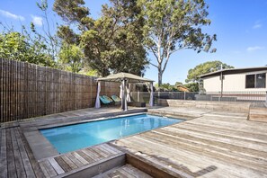 Relax in a serene backyard with a pool, deck, and shaded seating area surrounded by mature trees.