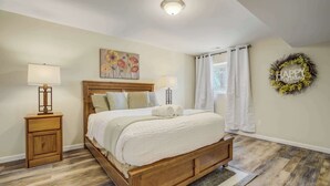 A bedroom with a wooden bed, two bedside tables with lamps, a floral painting, a wreath, and a window with curtains. - Iron View! Retreat w/ Hot Tub & Mountain Views (Colorado Springs)