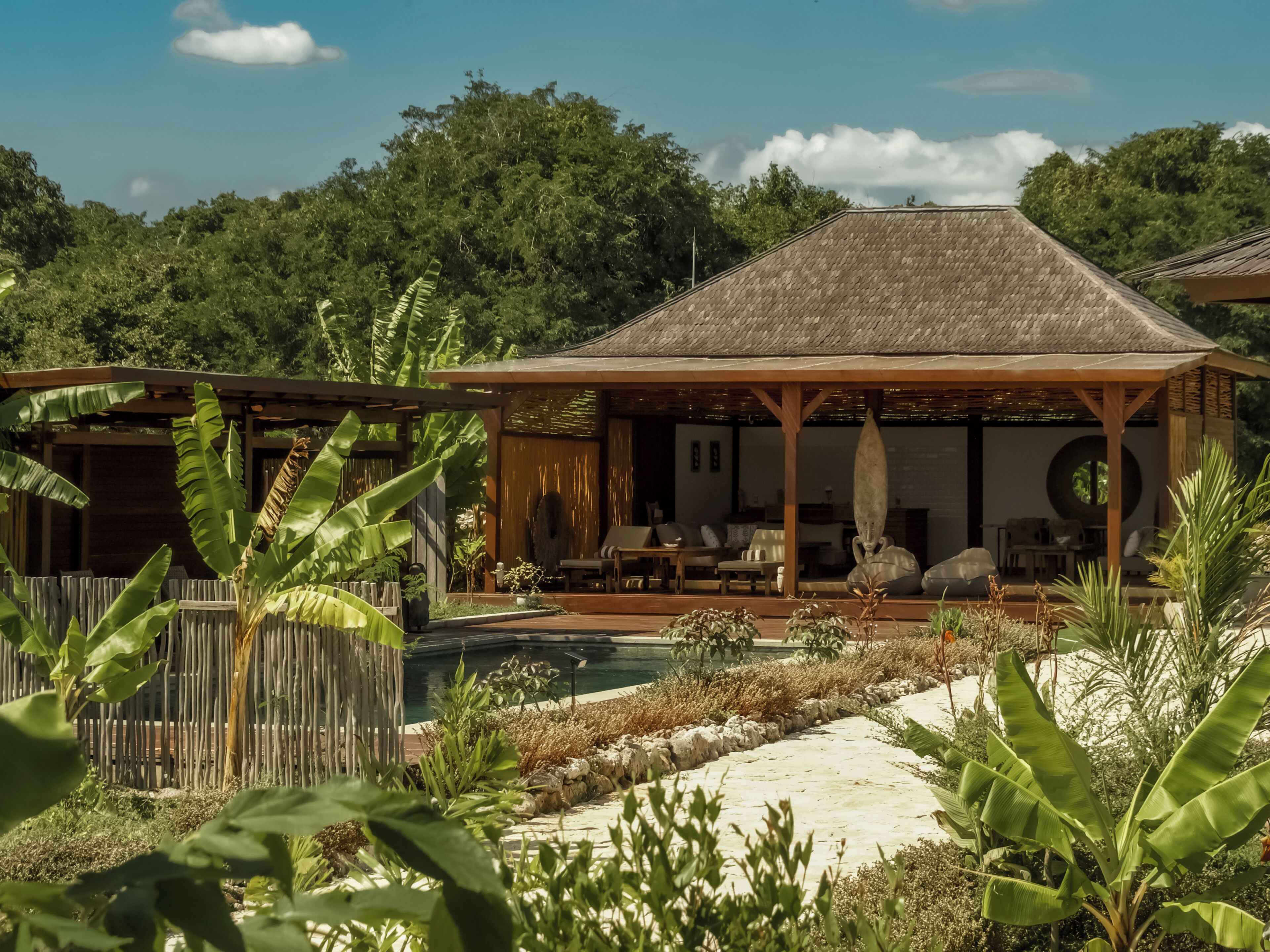 A bungalow with a thatched roof, a pool, and a wooden deck surrounded by tropical vegetation.