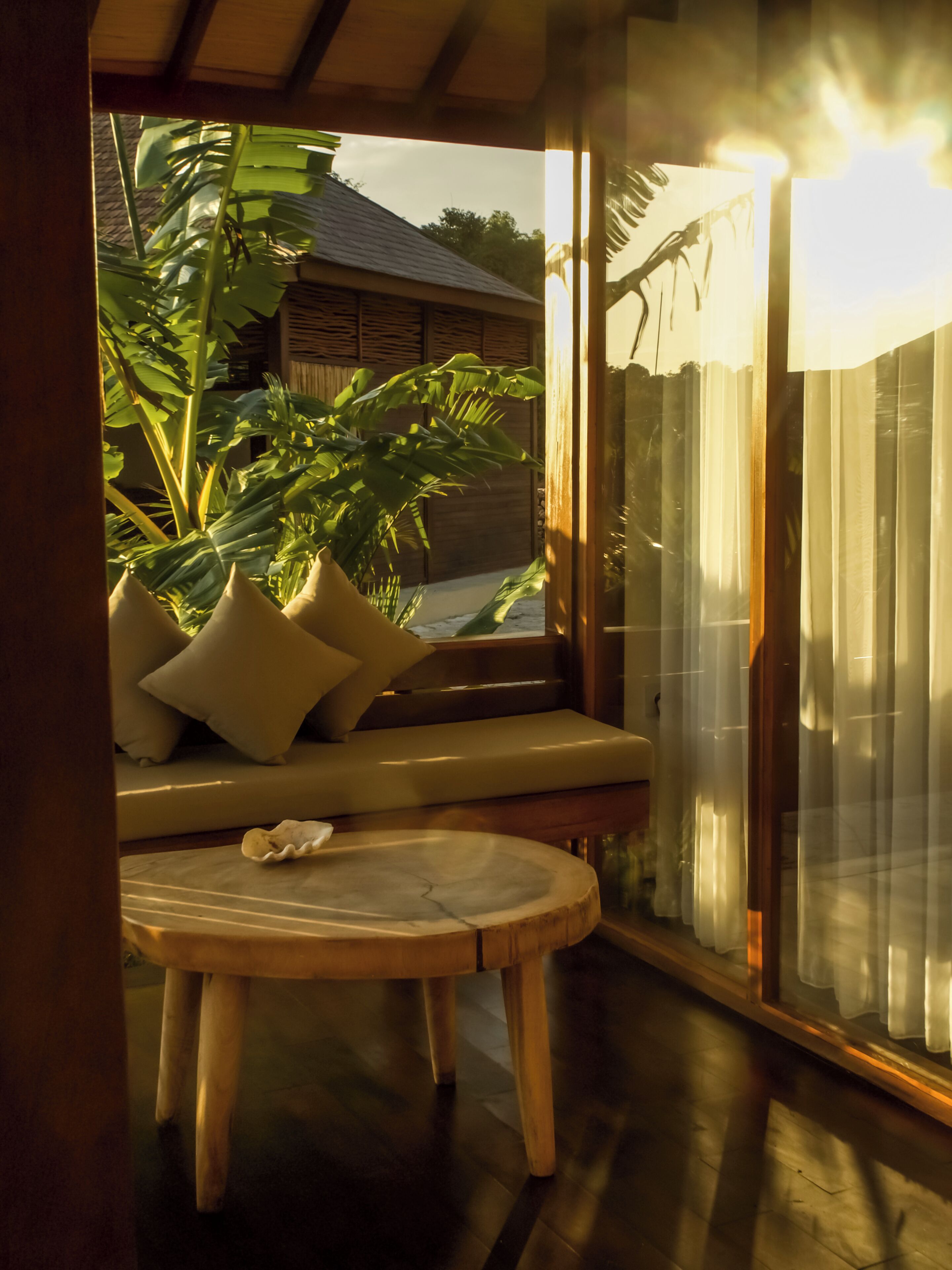 Ein Zimmer mit Holzboden, einem runden Holztisch, einer gepolsterten Bank mit Kissen und einem großen Fenster mit Vorhängen.