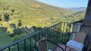 Varanda com cadeiras de vime e um banco acolchoado, com vista para uma paisagem montanhosa e uma estrada sinuosa.