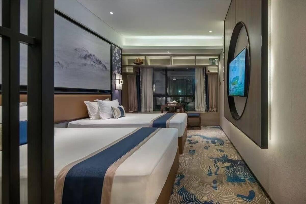 Ein Hotelzimmer mit zwei Betten, einem großen Fernseher, einem Gemälde und Blick auf die beleuchtete Stadt bei Nacht.