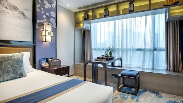 A hotel room with a large bed, a desk, a chair, a small table, a window with curtains, and a decorative ship model on the shelf. - Yuan Culture Hotel Shenzhen (Shenzhen)
