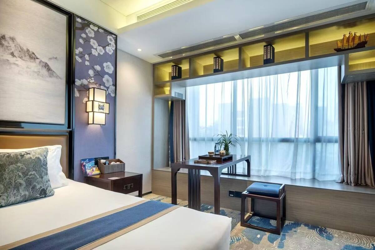 Ein Hotelzimmer mit einem großen Bett, einem Schreibtisch, einem Stuhl, einem kleinen Tisch, einem Fenster mit Vorhängen und einem dekorativen Schiffsmodell auf dem Regal.