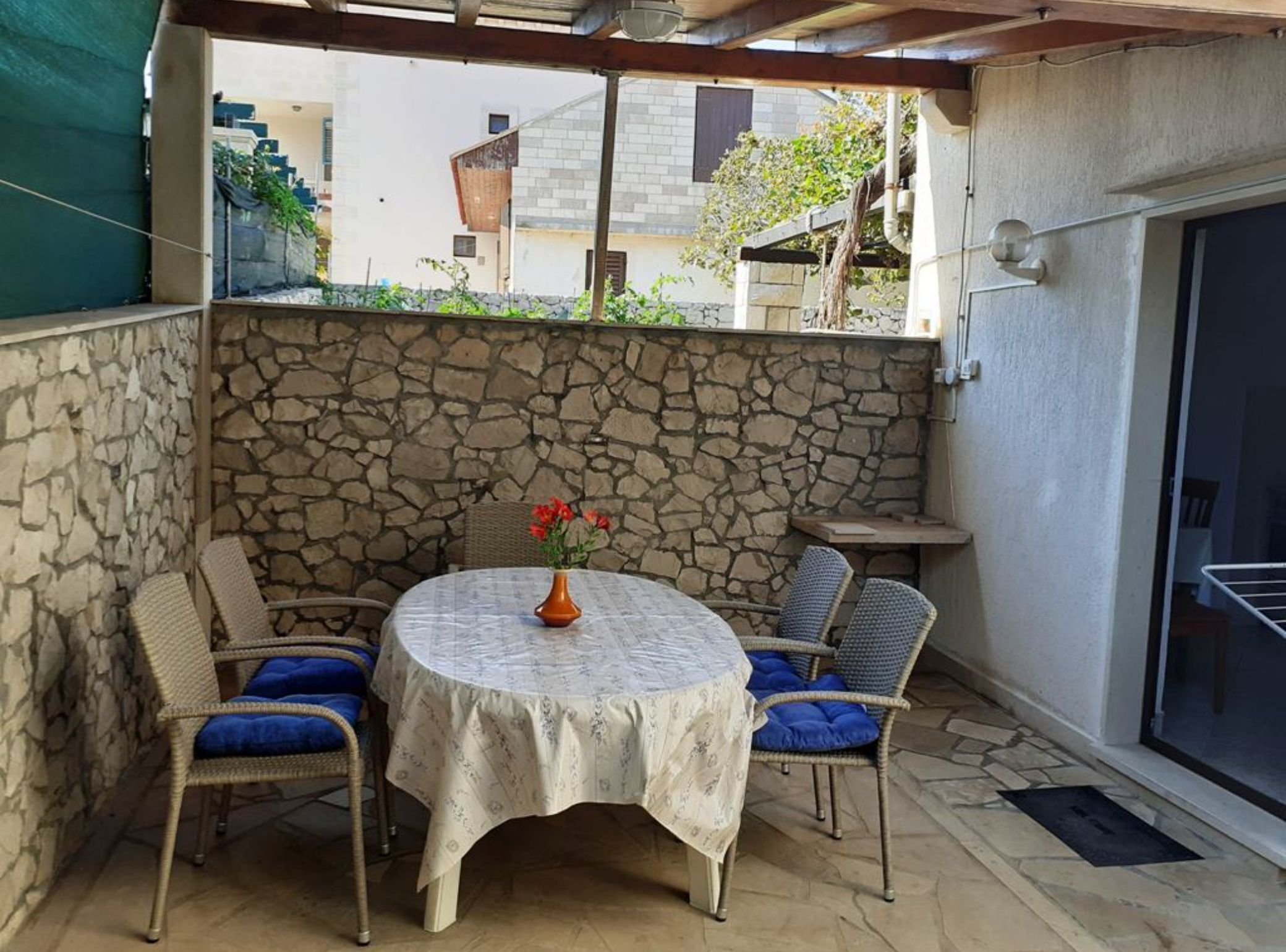 Un porche cubierto con una pared de piedra, una mesa redonda con cojines azules y un florero con flores.