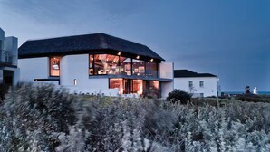A modern two-story house with large glass windows, a thatched roof, and a view of the sea.