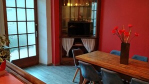 A dining area with a wooden table and chairs, a vase with red flowers, and a large window with frosted glass panels. - Gite la Rose des Alpes (Serres)