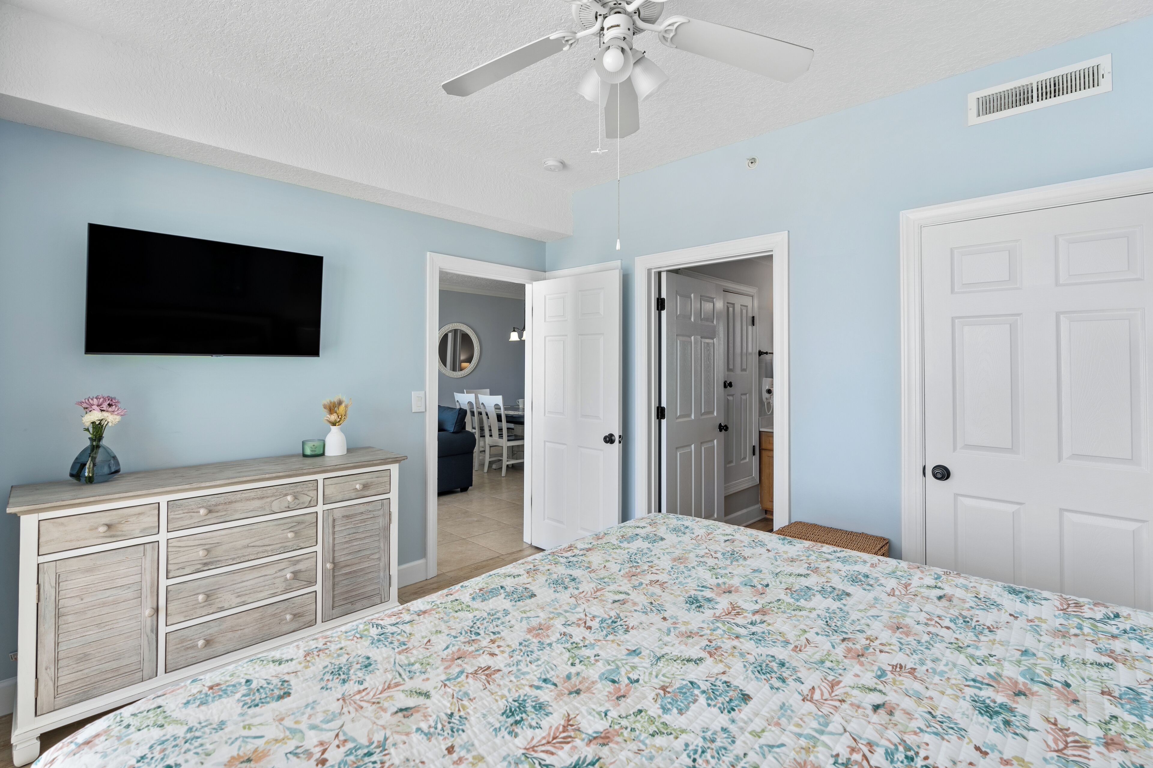 A bedroom with a bed, dresser, ceiling fan, and a TV on the wall.