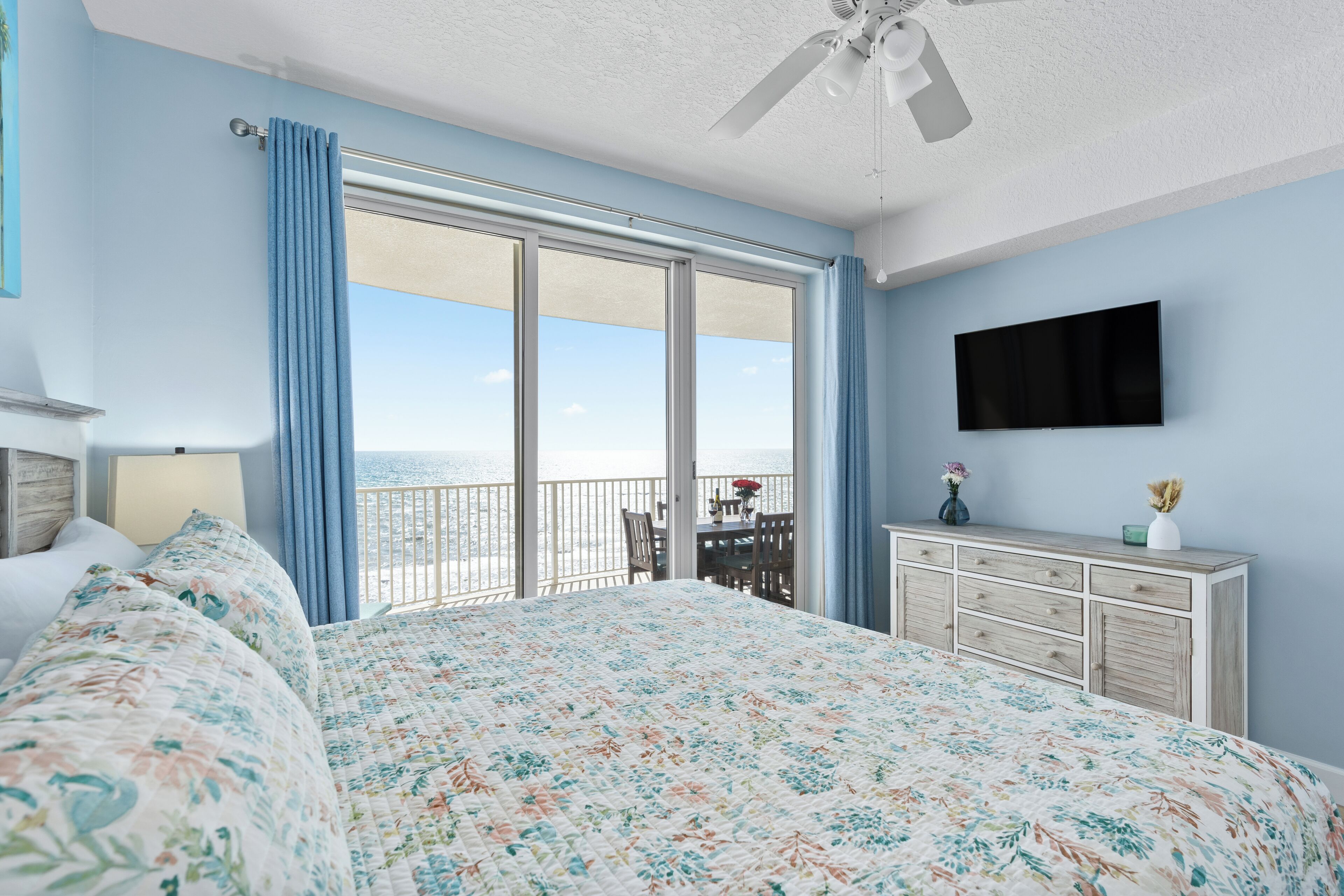A bedroom with a large bed, a dresser, a flat-screen TV, and a balcony with a view of the ocean.