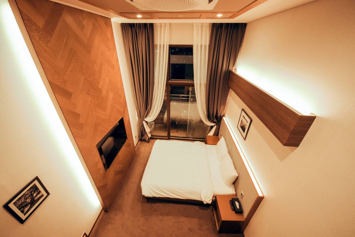 A hotel room with a bed, a bedside table, a nightstand, a framed picture, a window with curtains, and a view of the outside.