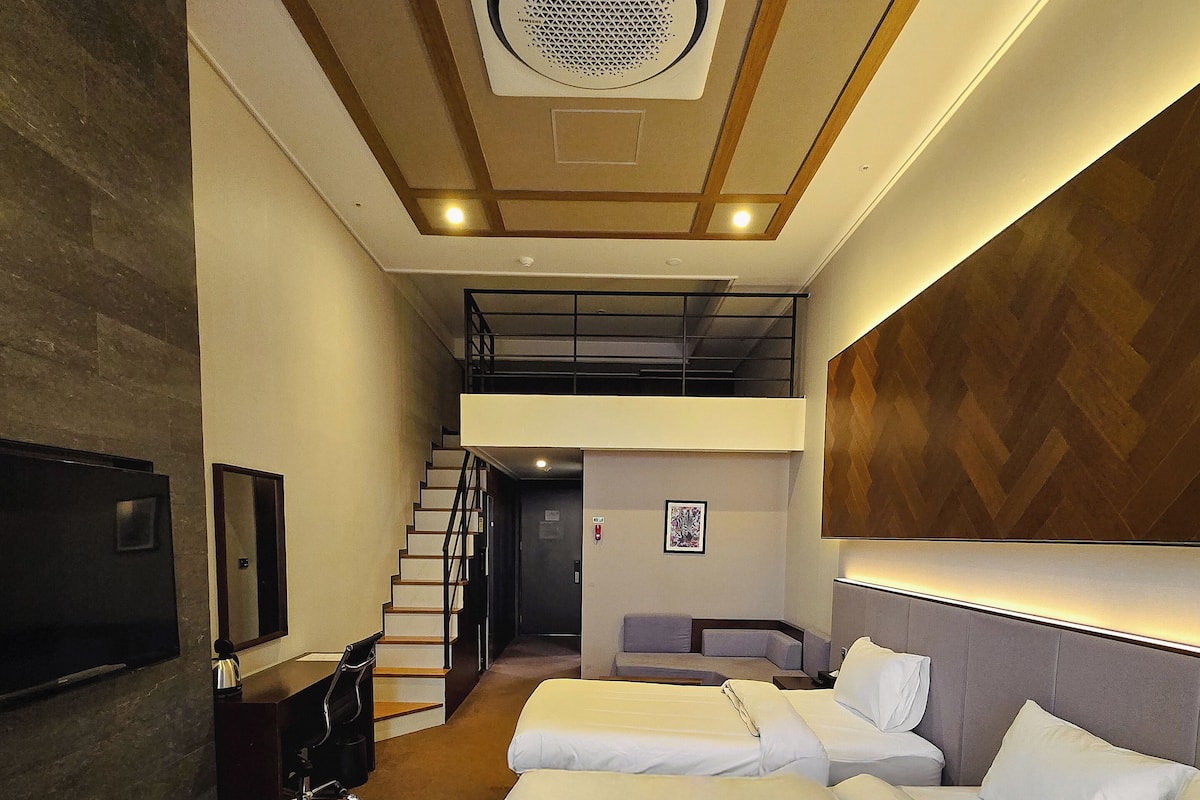 A hotel room with two beds, a TV, a desk, and a staircase leading to an upper level.