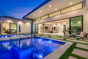 A modern outdoor pool area with a glass-enclosed patio, a dining area, and a well-lit interior space. - Indio Retreat • Pool + Hot Tub + Pickleball Court • Near La Quinta | Bravo (Indio)
