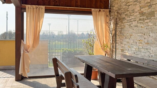 Un balcón de madera con una mesa y bancos, vista a un campo verde y un muro de piedra.