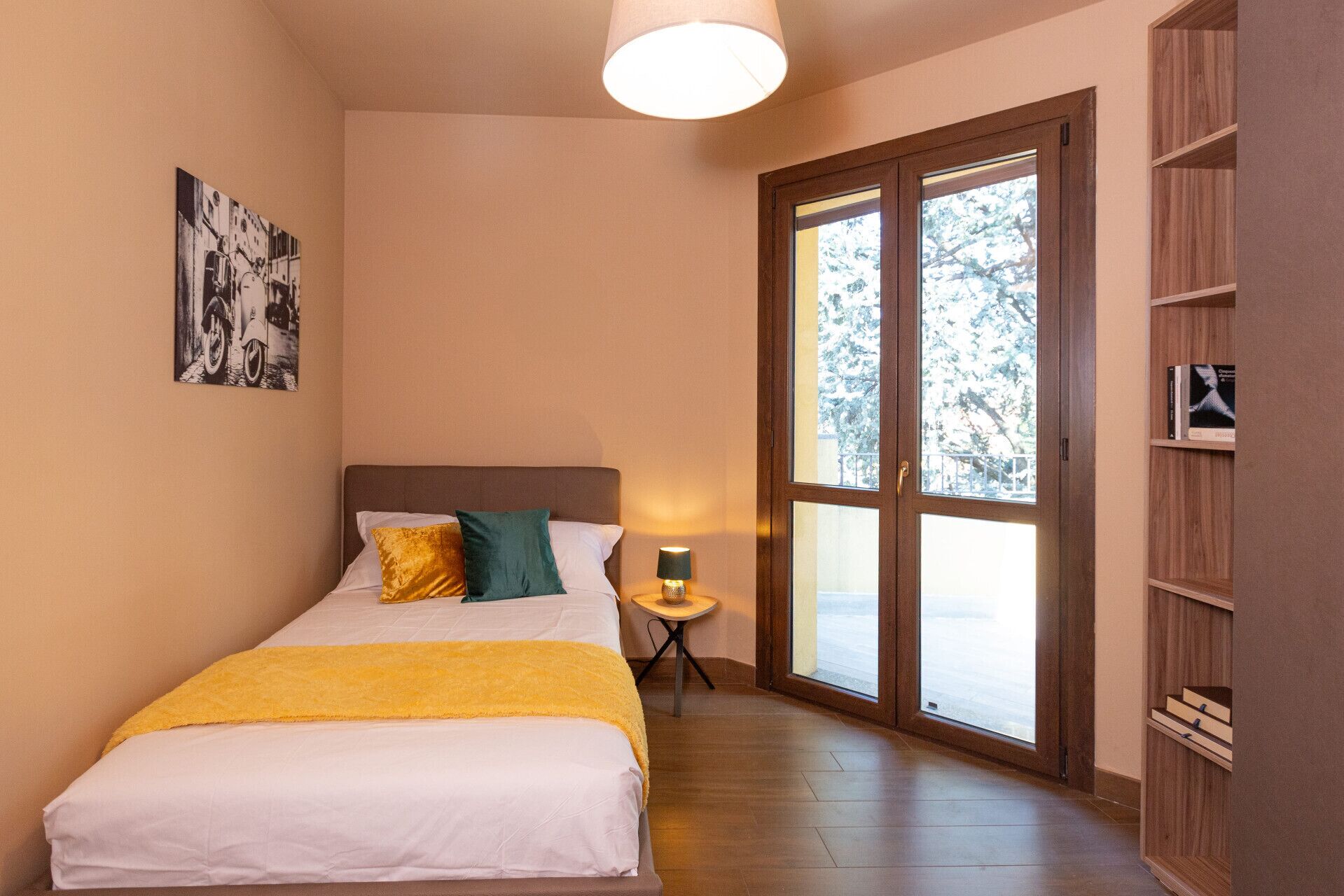 Quarto com cama, mesa de cabeceira, luminária, estante de livros e uma janela ampla com vista para árvores.