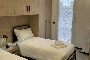 Cama bem arrumada com roupa de cama branca e uma toalha dobrada, luminária de cabeceira e um livro sobre mesa de cabeceira de madeira.