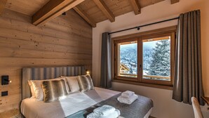 Dormitorio de una cabaña de madera con cama, almohadas, ventana con cortinas y vista a un paisaje nevado.