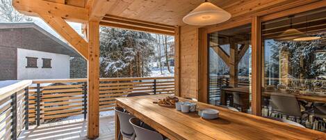 Una terraza de madera con zona de bar, asientos al aire libre y amplios ventanales con vistas a árboles cubiertos de nieve.