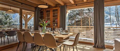 Un comedor con una mesa y sillas de madera, un florero con flores y vistas a árboles nevados a través de grandes ventanales.