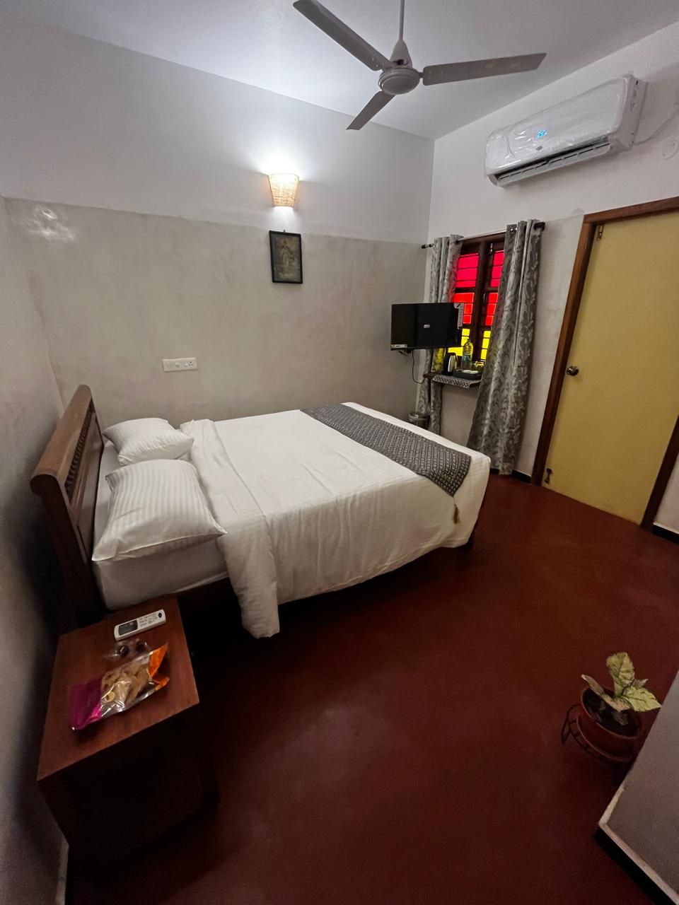 A hotel room with a bed, a bedside table with a remote control, a ceiling fan, an air conditioning unit, a TV on a stand, a door, and a potted plant.