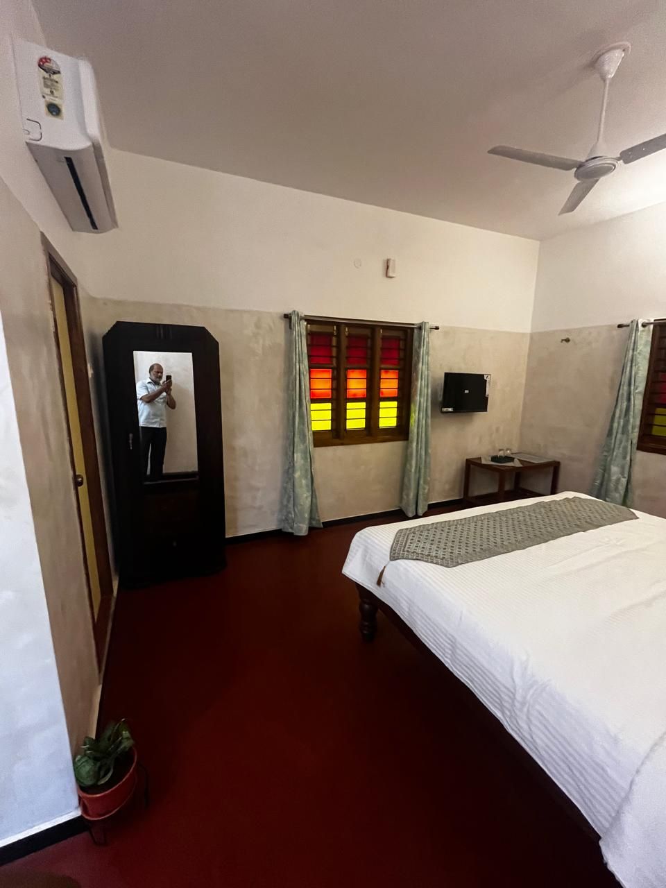 Ein Hotelzimmer mit einem Bett, einem Deckenventilator, einem Fenster mit Vorhängen, einem kleinen Tisch, einem Fernseher und einer Topfpflanze.