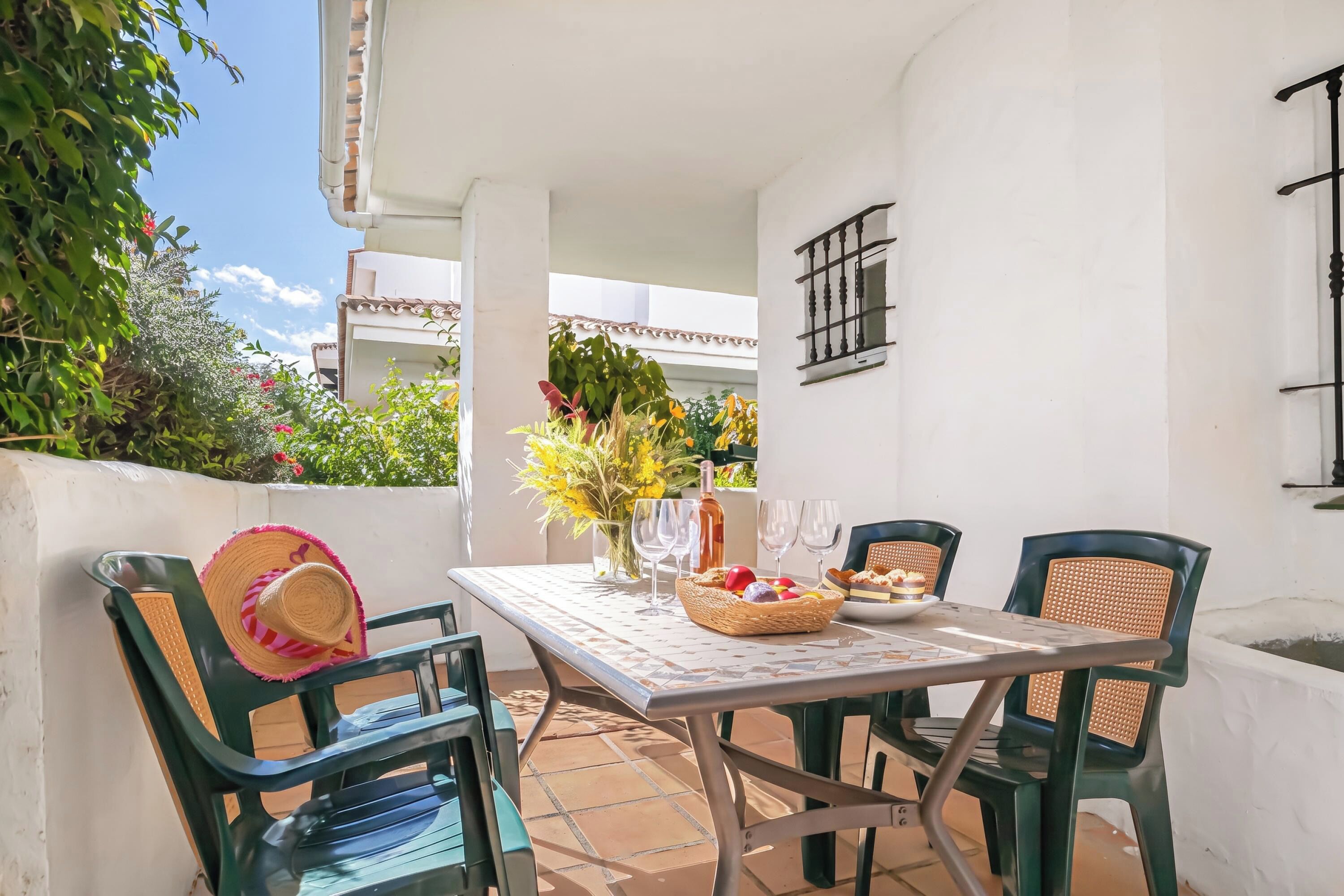 An outdoor patio with a table set for two, surrounded by green chairs, a vase with flowers, and a bottle.