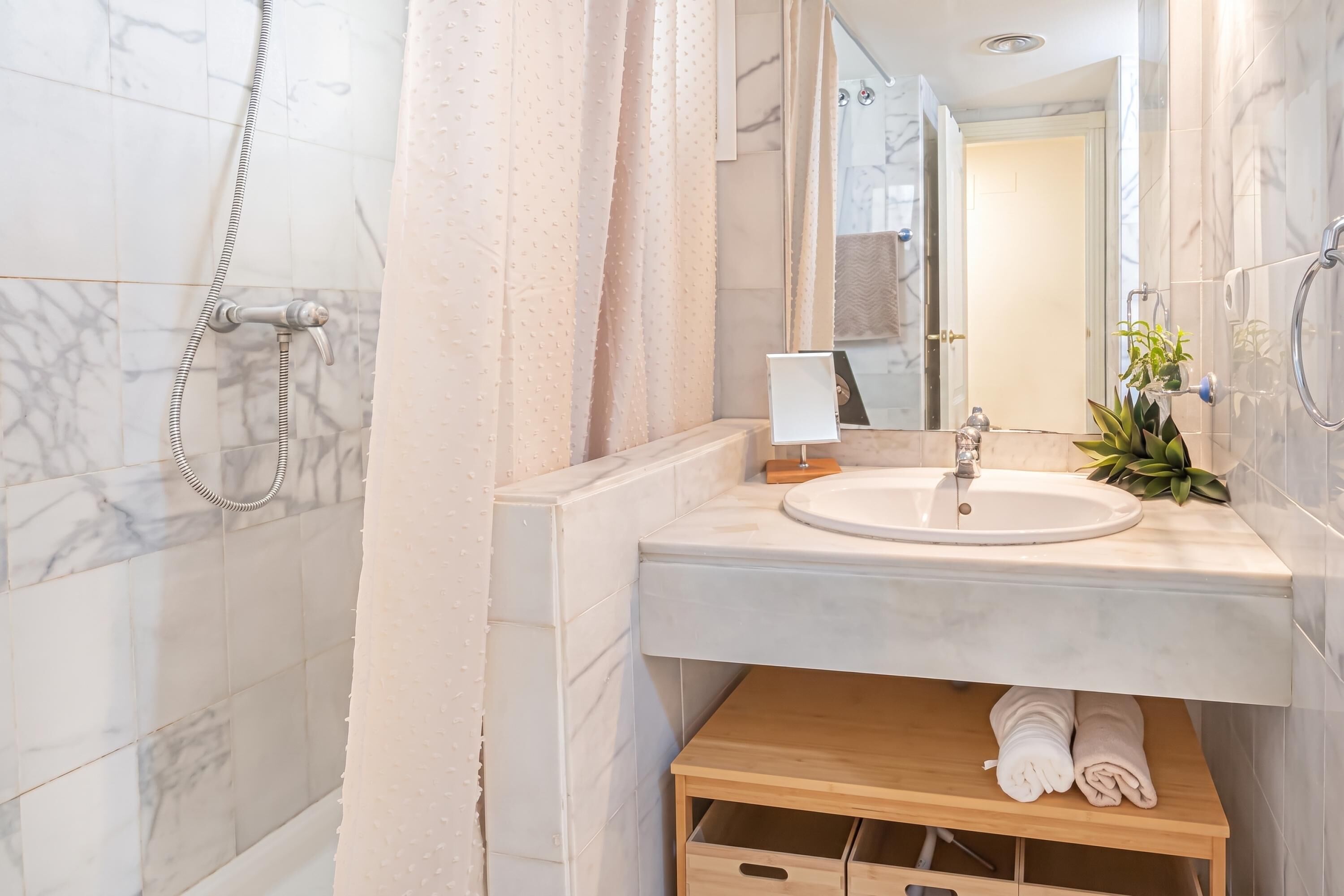 Ein Badezimmer mit einer Marmor-Fliesenwand, einer Dusche inklusive Handbrause, einem Waschbecken mit Spiegel darüber und einem Holzregal mit Handtüchern.