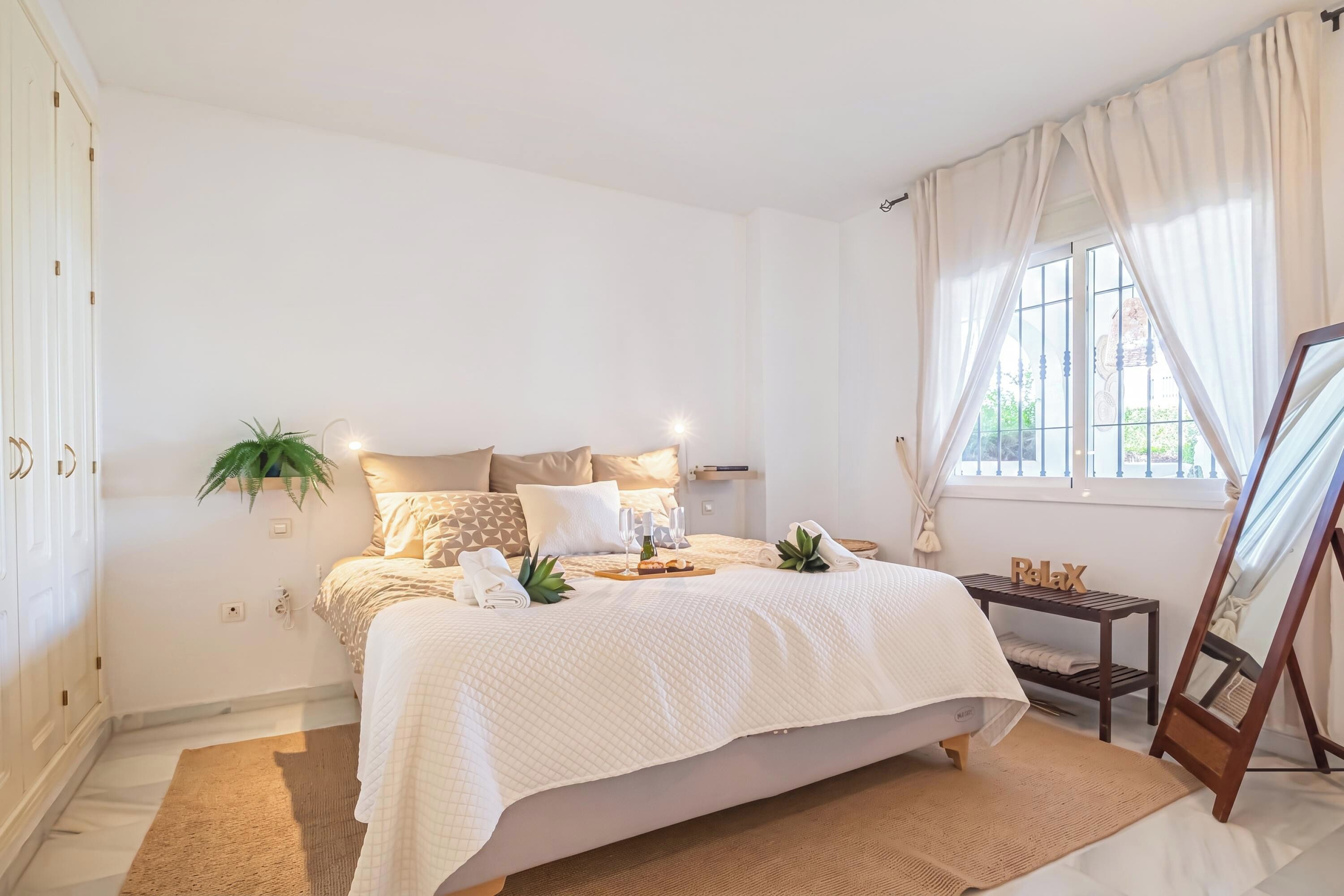 A well-lit bedroom with a bed, bedside table, and a window with curtains.
