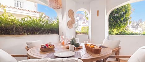 A round table with white chairs, a bottle of wine, glasses, and a fruit basket.
