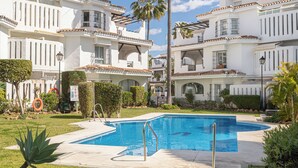 Una piscina rodeada de edificios blancos con balcones, cielo despejado y vegetación.