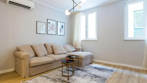 A modern living room with a beige sectional sofa, a small side table, a patterned rug, and a ceiling light fixture. - Cultural Haven Central Two Bedroom Apartment in Zadar With City Views (Zadar)