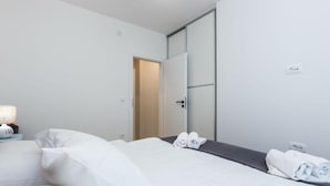A hotel room with a bed, a nightstand, a lamp, and a door leading to another room. - Cultural Haven Central Two Bedroom Apartment in Zadar With City Views (Zadar)