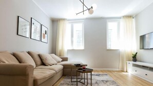 A modern living room with a beige sofa, a coffee table, and a TV on a white cabinet. There are two windows with curtains, a hanging light fixture, and framed pictures on the wall. - Cultural Haven Central Two Bedroom Apartment in Zadar With City Views (Zadar)
