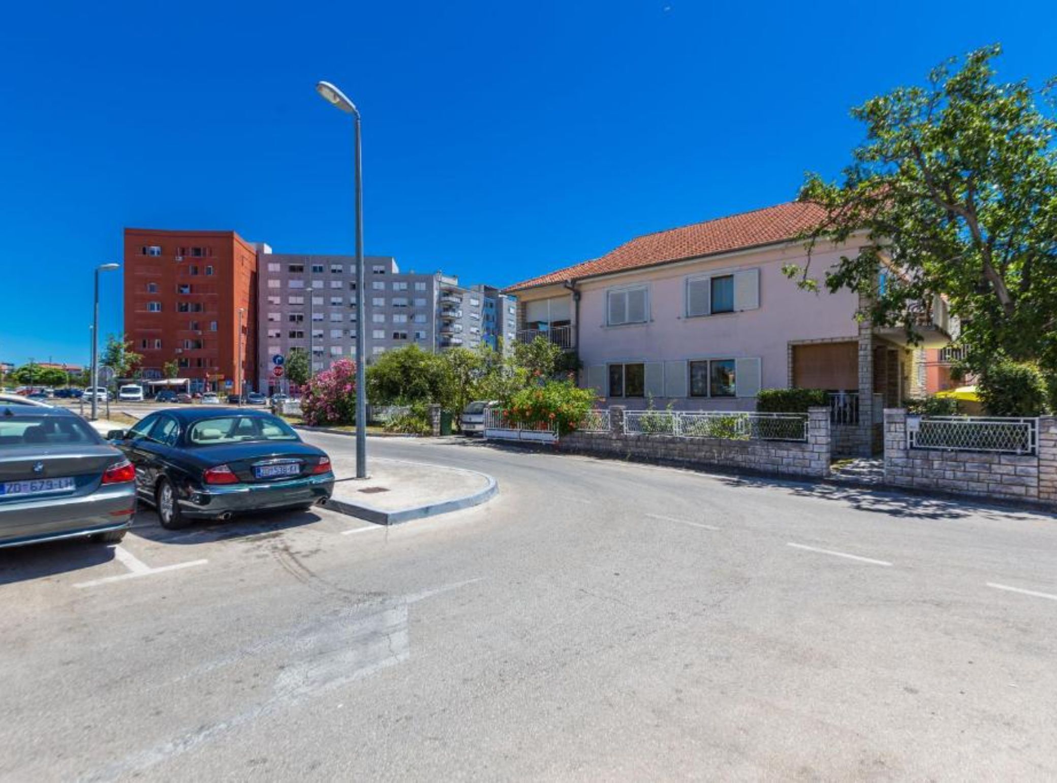 Une vue de rue avec un espace de stationnement, un bâtiment en briques rouges et une maison rose avec un jardin.