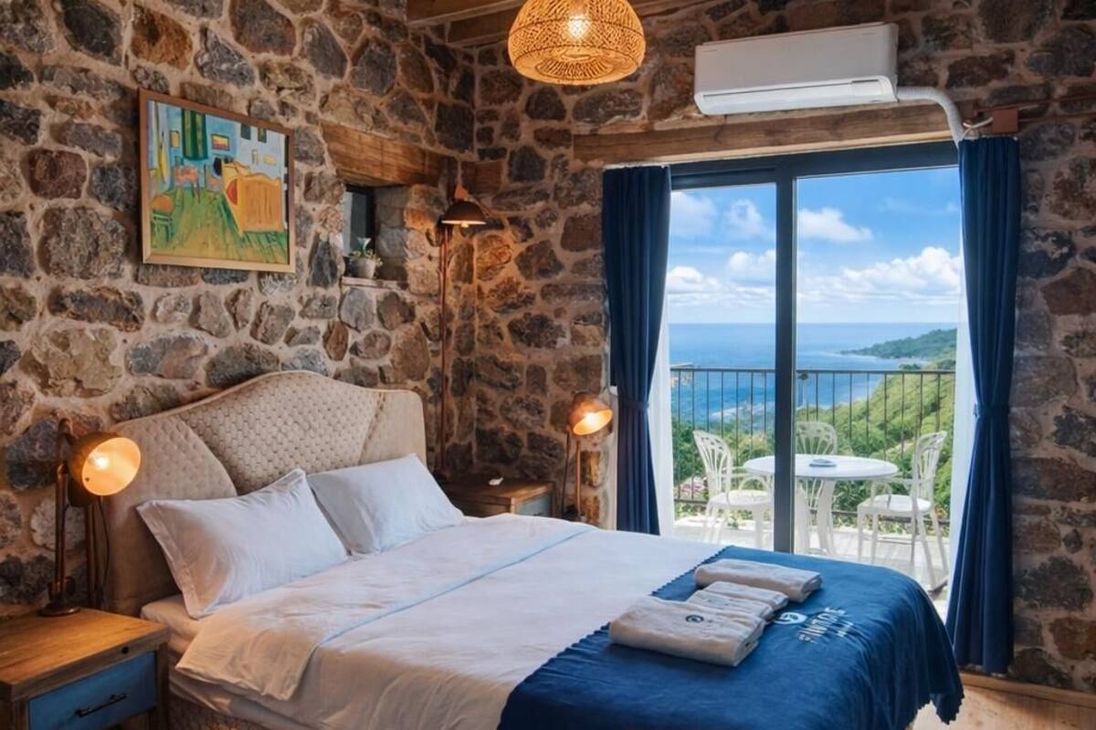 A bedroom with a stone wall, wooden ceiling beams, a bed with a blue bedspread, a painting, and a view of the sea.