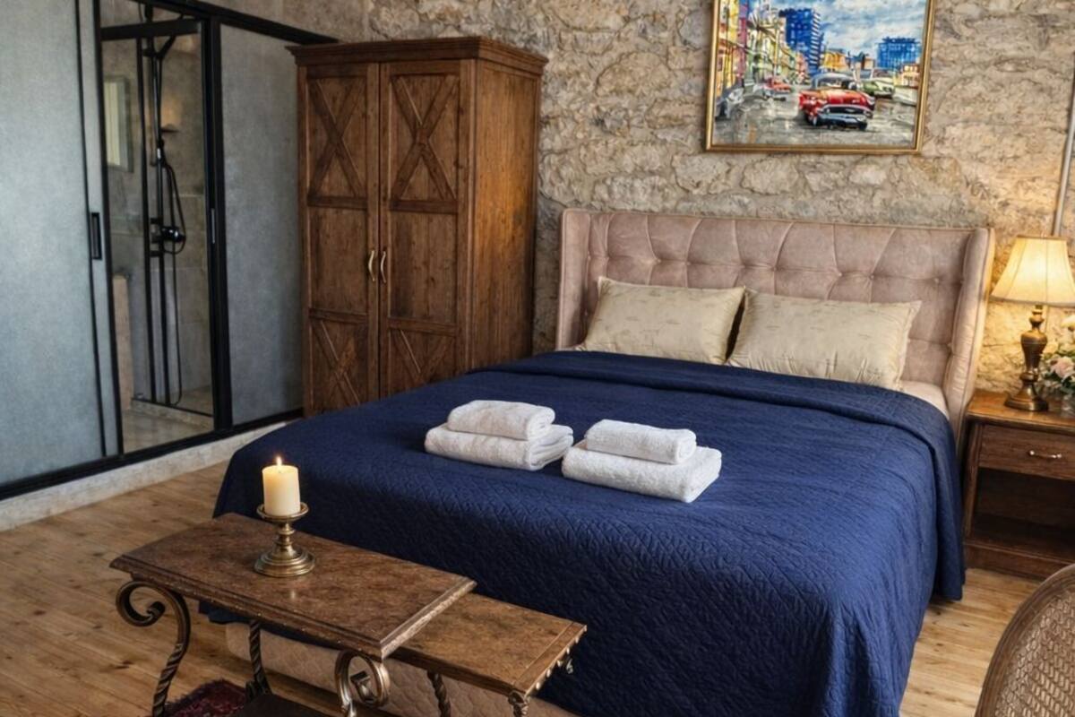 A bedroom with a stone wall, a bed with blue bedding, a wooden cabinet, a chair, and a small table with a candle.