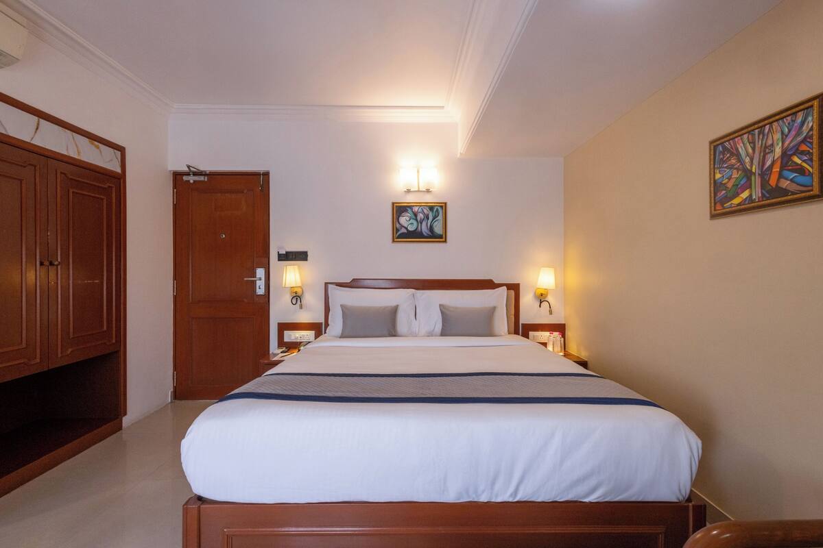 A hotel room with a large bed, wooden wardrobe, two bedside lamps, a framed artwork, and a door.