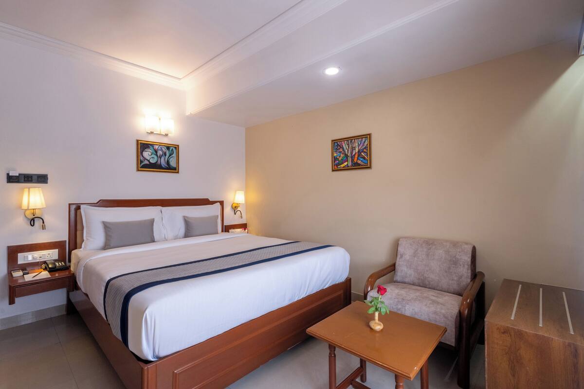 A hotel room with a large bed, two bedside tables with lamps, a small wooden table with a vase, a chair, and two wall-mounted artworks.