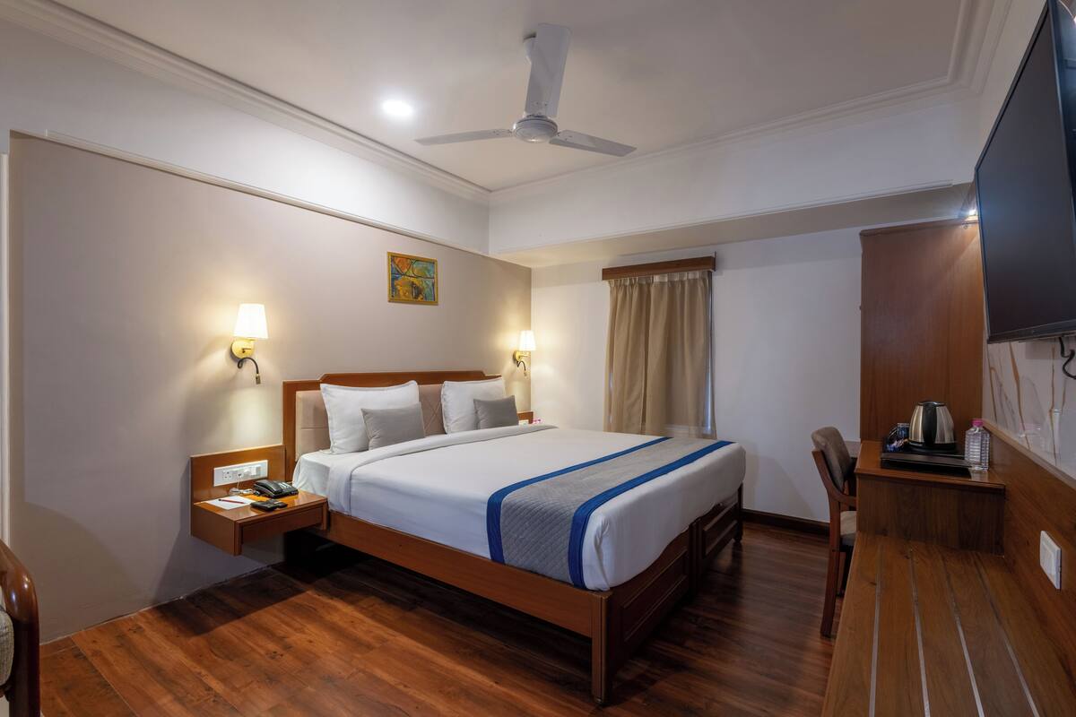 A hotel room with a wooden bed, bedside tables, a ceiling fan, a flat-screen TV, and a window with curtains.