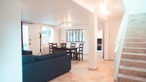 Amplia sala de estar con sofá, mesa de comedor y sillas. Se ve una escalera a la derecha.
