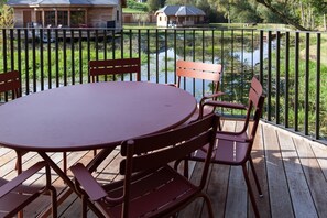 A round table with four chairs on a wooden deck, overlooking a calm body of water with houses in the background. - Cozy Cabins in Vencimont (Vencimont)