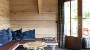 A wooden interior with a bench, cushions, a round wicker table, and a view through a window with curtains. - Cabin in Vencimont near Ardennes Trails (Vencimont)