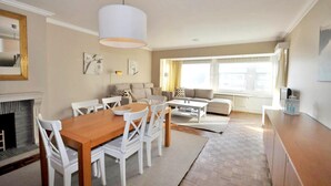 Ein modernes Wohnzimmer mit Essbereich, ausgestattet mit einem Holztisch, weißen Stühlen, einer Couch und einem großen Fenster mit Vorhängen.