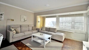 Ein modernes Wohnzimmer mit einem beigen Sofa, einem weißen Couchtisch und einem großen Fenster mit Blick auf Gebäude.